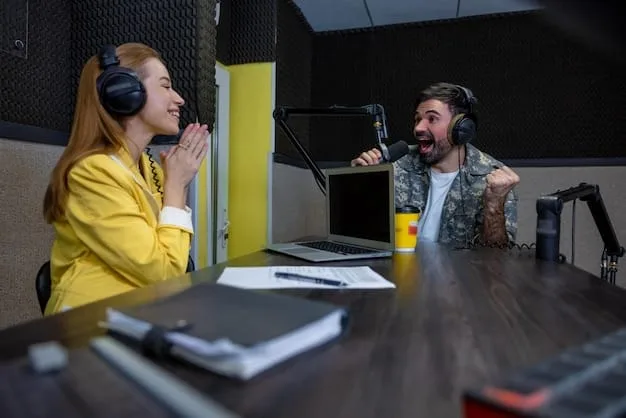 A brightly lit studio during a live recording of a comedy podcast. Two comedians are sitting at a table with microphones, laughing. There are visible audience members in the background, also laughing.