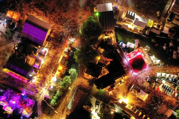 Aerial view of New Orleans during Jazz Fest, showing the fairgrounds packed with stages, tents, food vendors, and a vibrant, diverse crowd enjoying the music and atmosphere.